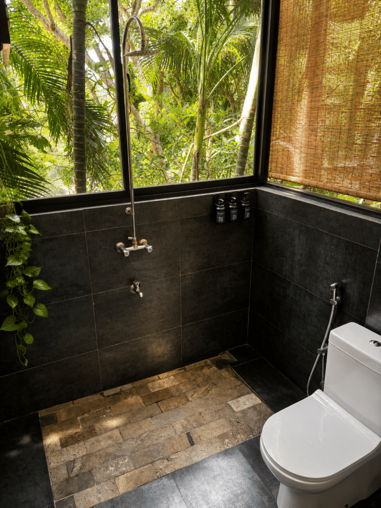 Open-air bathroom with black tiling, rainfall shower and toilet surrounded by tropical greenery at Salt House, Hiriketiya Bay, Sri Lanka